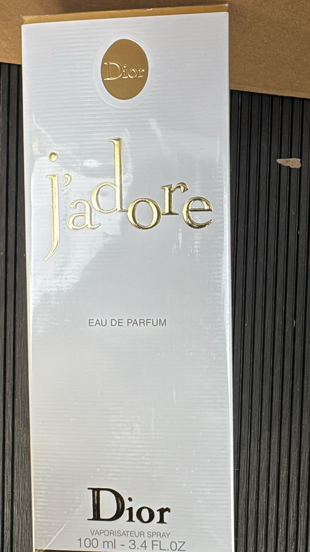 Dior J'adore Eau de Parfum 100ml box packaging, featuring a white textured background with gold foil lettering and a gold circular Dior logo at the top.