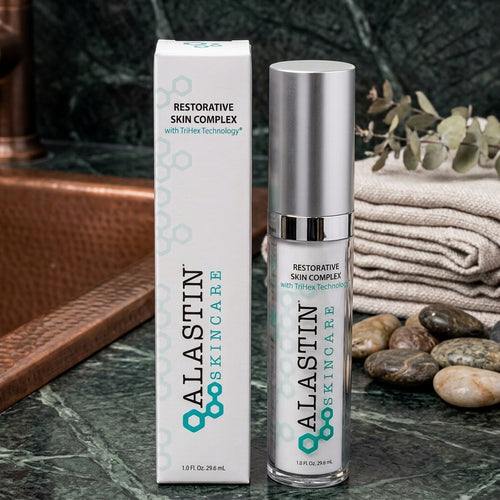 Alastin Skincare Restorative Skin Complex shown with its white and teal outer box and the clear cylindrical bottle with a silver cap, styled on a dark marble surface with folded fabric, stones, and greenery in the background.