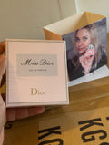A hand holds a square Dior Miss Dior Eau de Parfum box in the foreground, with an open Dior-branded inner box and printed insert behind it on a cardboard surface.