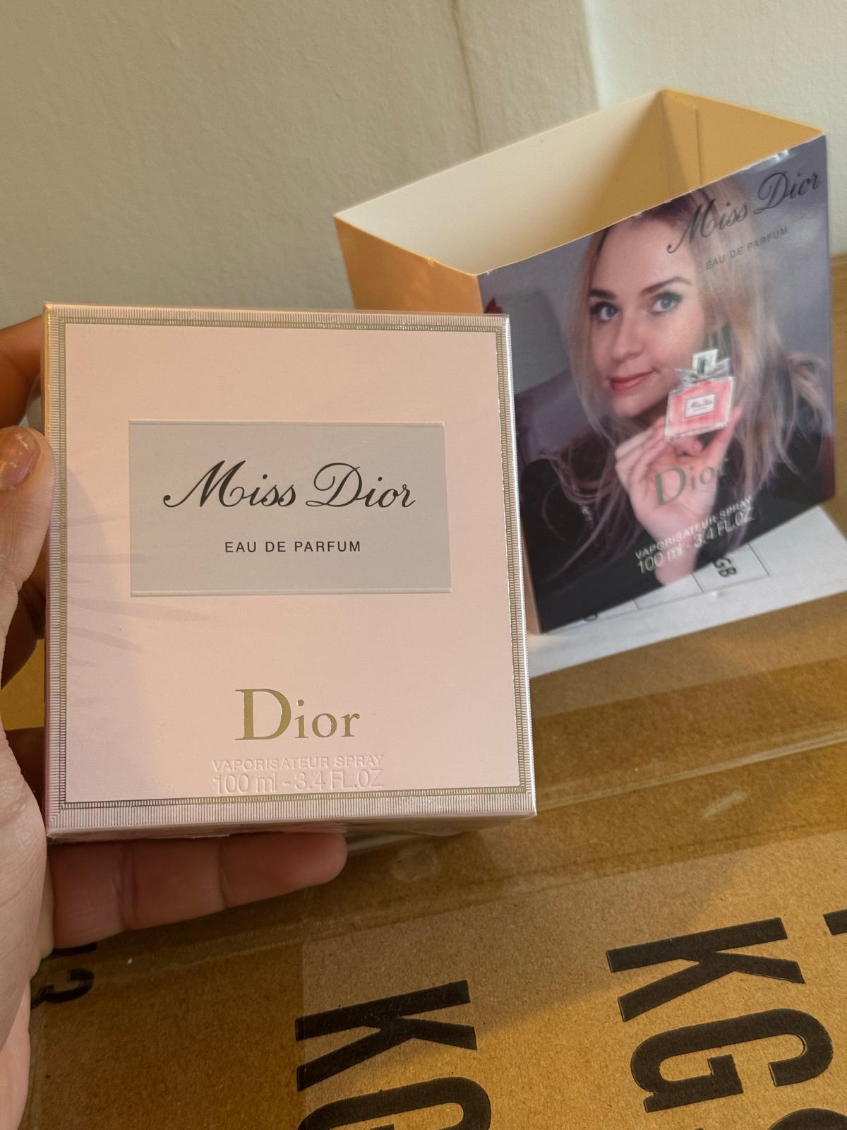 A hand holds a square Dior Miss Dior Eau de Parfum box in the foreground, with an open Dior-branded inner box and printed insert behind it on a cardboard surface.