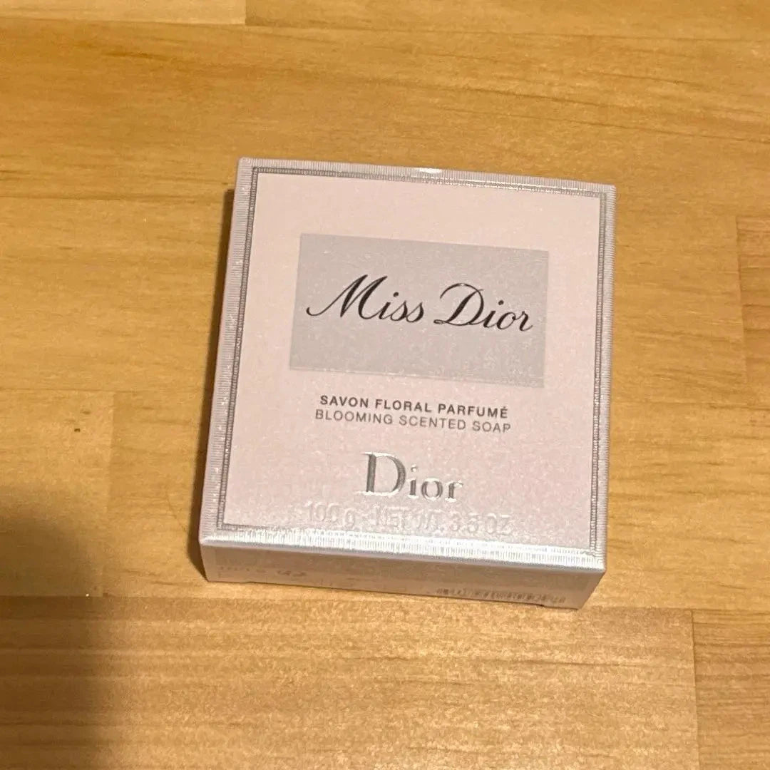 A square box of Dior Miss Dior Blooming Scented Soap placed on a light wood surface. The box is pale pink with silver-gray text and border details.