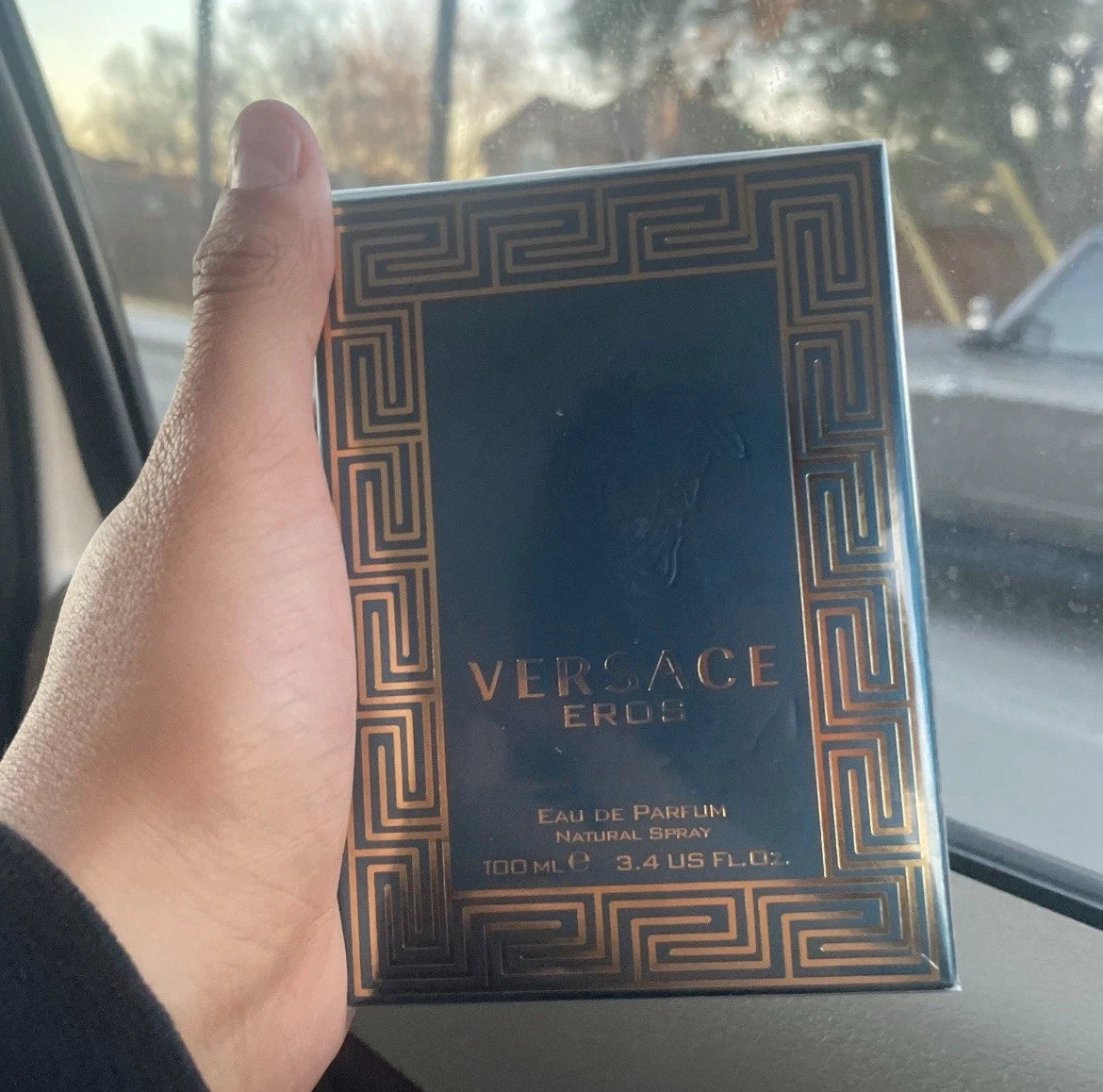 A hand holding a box of Versace Eros Eau de Parfum in a car, with the black box featuring a gold Greek key pattern border and gold lettering.