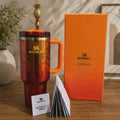 Stanley Karolo 32 oz chevron stainless steel tumbler in a red and orange gradient finish, shown with its orange packaging box and branded card on a wooden table.