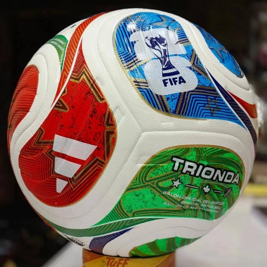 A close-up of a FIFA-branded soccer ball with white panels and large red, blue, green, and gold graphic sections. The ball is photographed indoors against a dark blurred background, with a small yellow stand visible at the bottom.