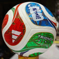 A close-up of a FIFA-branded soccer ball with white panels and large red, blue, green, and gold graphic sections. The ball is photographed indoors against a dark blurred background, with a small yellow stand visible at the bottom.