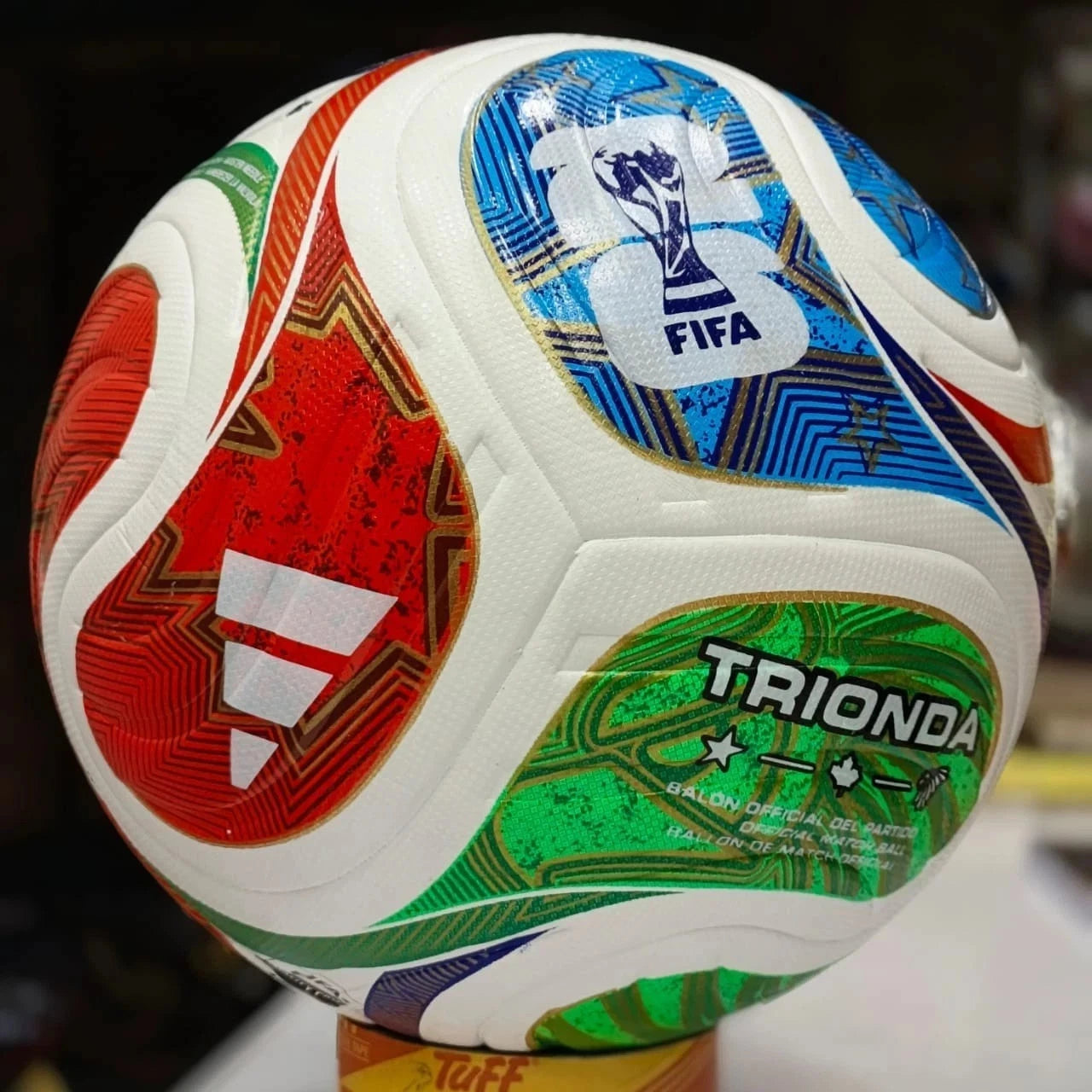 A close-up of a FIFA-branded soccer ball with white panels and large red, blue, green, and gold graphic sections. The ball is photographed indoors against a dark blurred background, with a small yellow stand visible at the bottom.