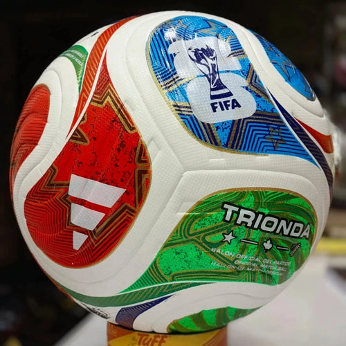A close-up of a FIFA-branded soccer ball with white panels and large red, blue, green, and gold graphic sections. The ball is photographed indoors against a dark blurred background, with a small yellow stand visible at the bottom.
