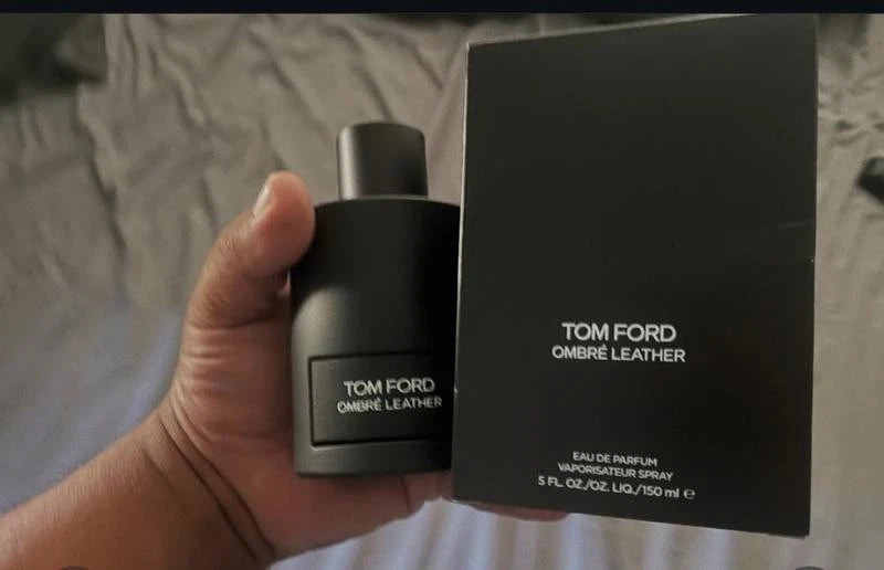 A hand holding a black Tom Ford Ombre Leather Eau de Parfum bottle next to its black packaging box, with a wrinkled gray fabric background.