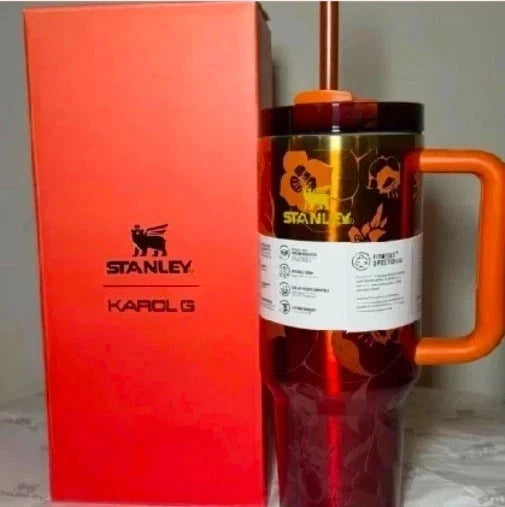 A Stanley Karol G tumbler with an orange handle and straw, standing next to its bright orange retail box. The tumbler features a black and orange floral pattern with the Stanley logo.