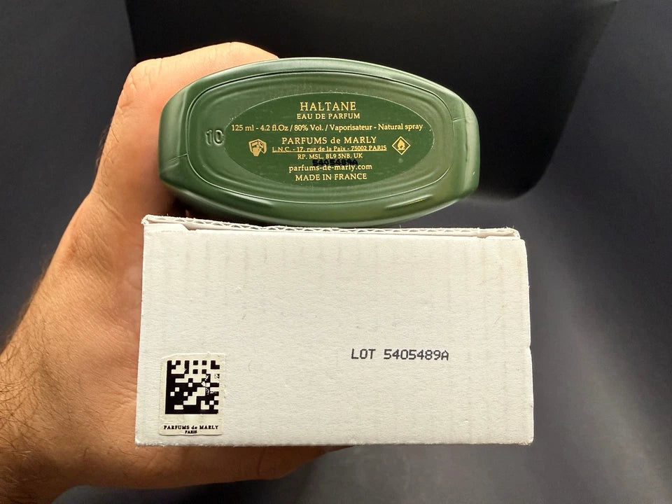 A hand holding a green bottle of Halthane Eau de Parfum above its white cardboard packaging box, with the brand name and product details visible on the bottle.