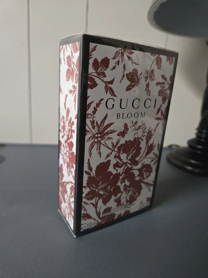 Gucci Bloom perfume box with a white background and dark red floral pattern, standing on a dark gray surface.