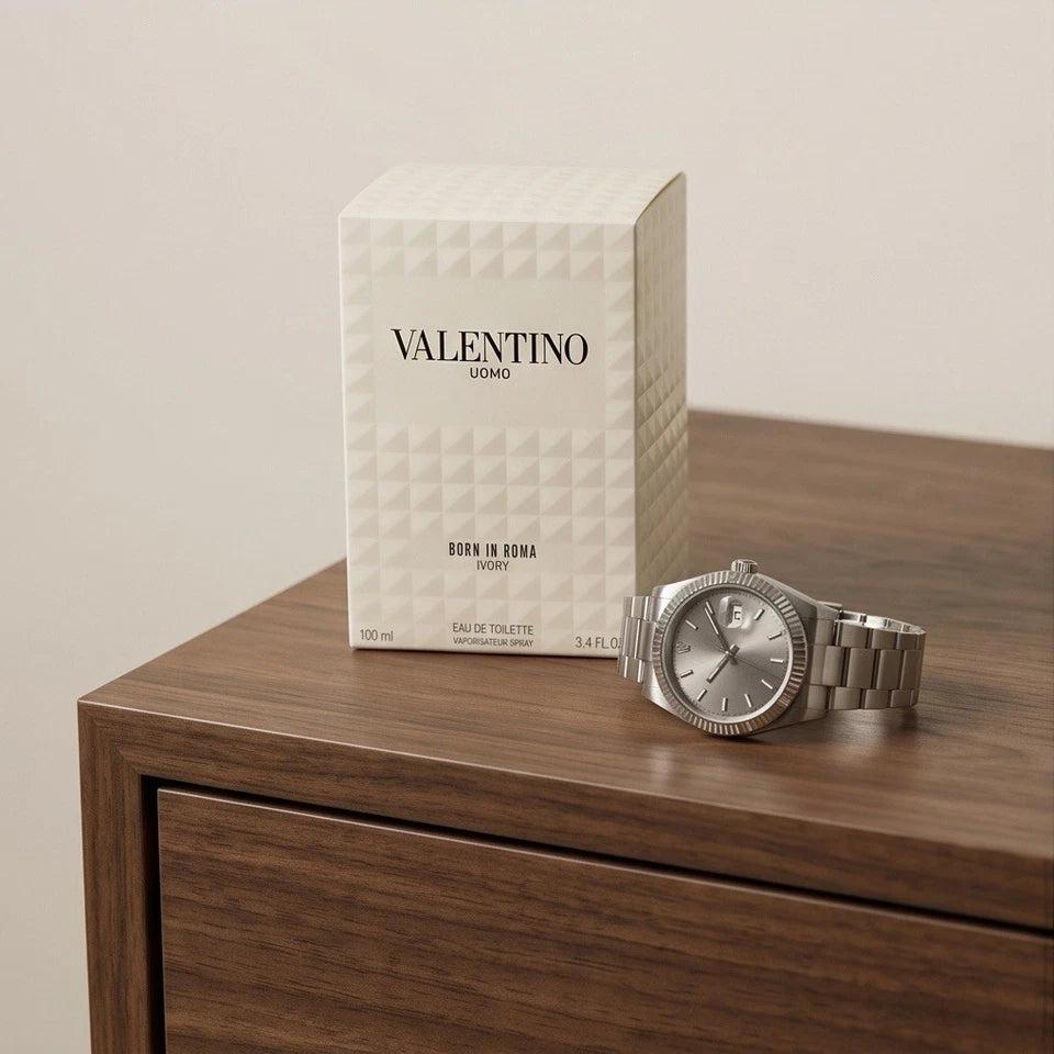 A silver-toned Valentino Uomo wristwatch with a metal link bracelet sits on a dark wood table next to its textured off-white retail box, set against a neutral beige background.