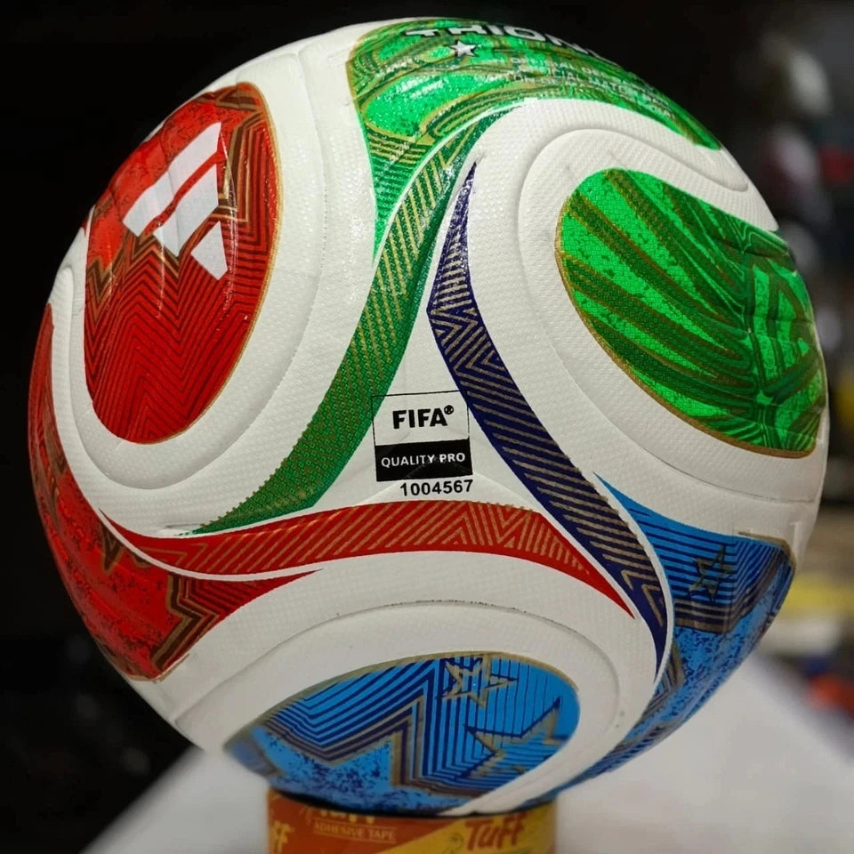 A close-up of a FIFA-branded soccer ball with a white base and large red, green, blue, and dark navy graphic panels, photographed against a blurred dark background.
