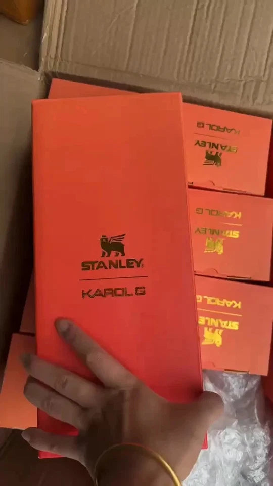 A hand holds a bright orange Stanley Karol G shoebox in front of several stacked orange boxes inside a cardboard shipping box. The front box features the black Stanley lion logo and 'STANLEY KAROL G' text.