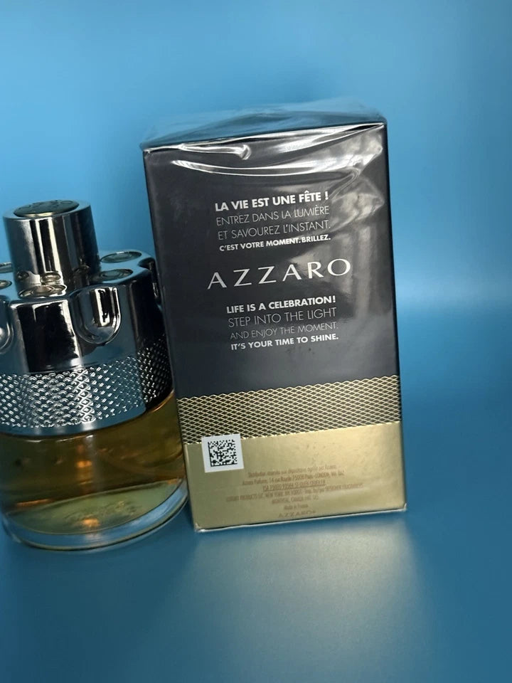 A bottle of Azzaro fragrance next to its original packaging box, both featuring the brand name 'AZZARO' in white text. The bottle has a textured silver cap and base with amber-colored liquid inside, while the box is black with a metallic gold lower section.