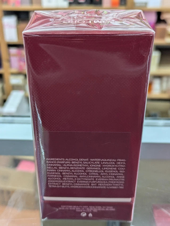 A rectangular perfume box in deep burgundy with a glossy finish, wrapped in clear protective plastic. The back of the box features small white text, and the background shows a blurred store shelf environment.