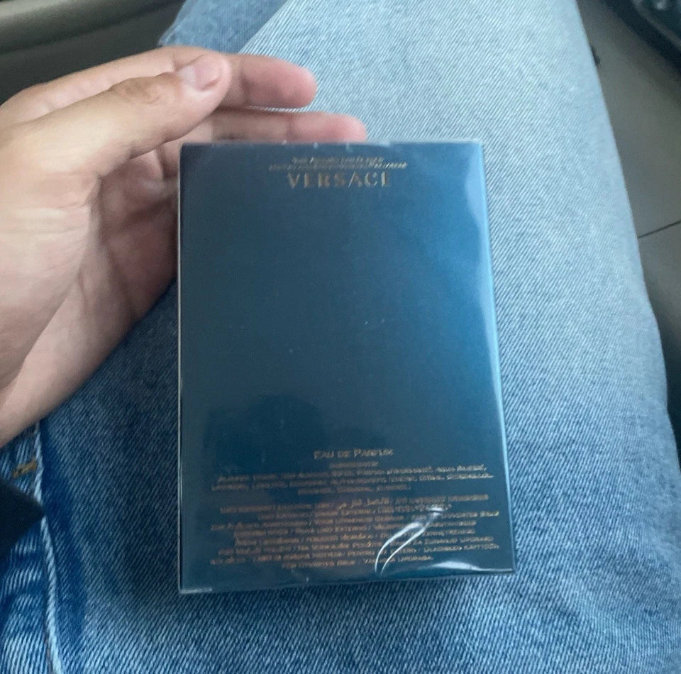A hand holding a square Versace fragrance box wrapped in clear plastic, set against blue denim fabric.