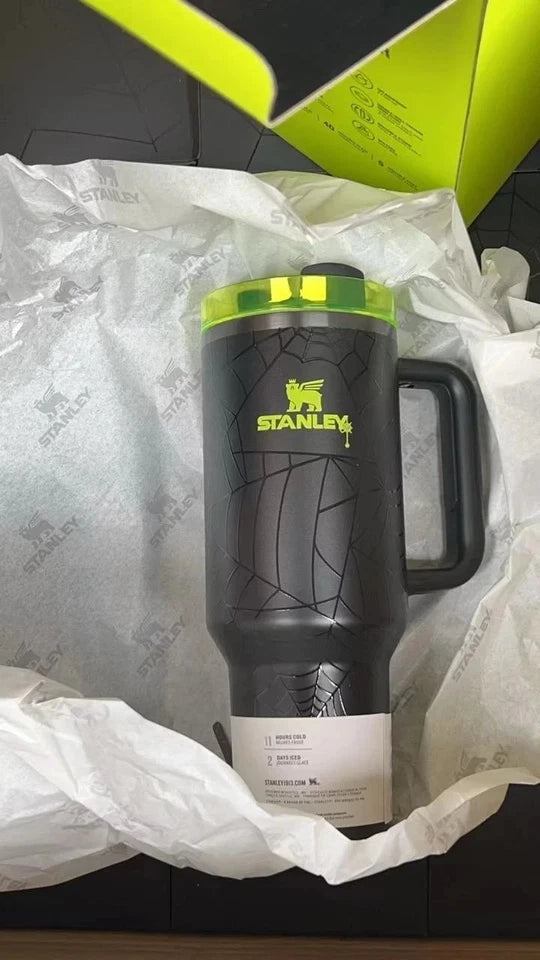 A black Stanley insulated tumbler with a bright green lid and green logo, nestled in white tissue paper with a green Stanley box visible in the background.